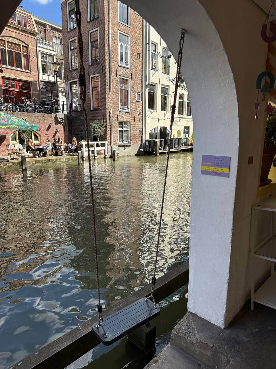 A wooden swing hanging under a canal-side archway in Utrecht, with a café and houseboats in the background