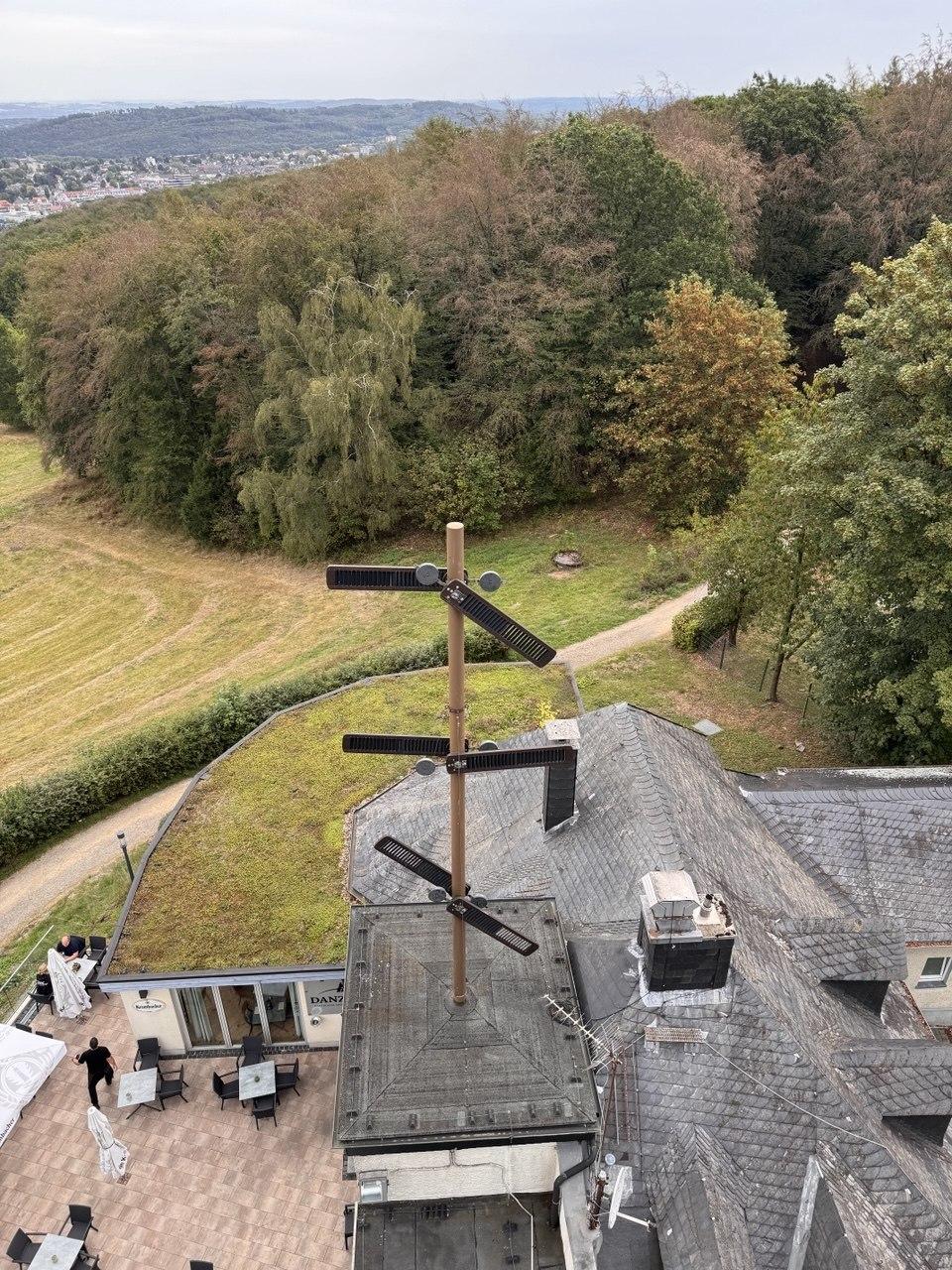 Aerial view from the Danzturm tower looking down at a telegraph-style antenna mast on the roof, with the restaurant terrace and Sauerland hills below